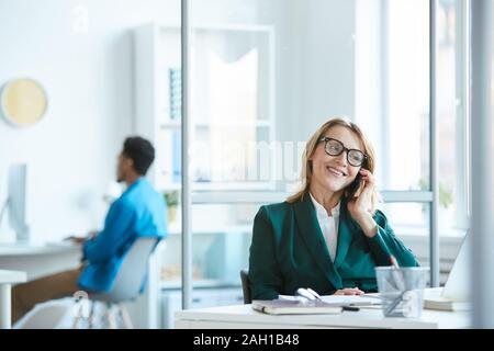 Coppia manager in occhiali seduta al tavolo parlando al telefono cellulare e sorridente in ufficio Foto Stock