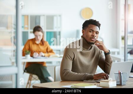 Ritratto di giovane africano lavoratore di ufficio guardando la fotocamera con pensieroso vista mentre seduto al suo posto di lavoro con il computer portatile Foto Stock