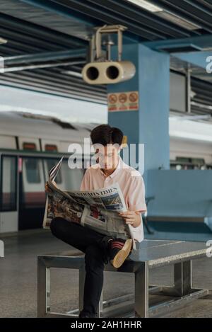 Uomo singaporiano in camicia rosa che legge gli Straits Times alla stazione MRT di Queenstown con MRT Train in background, Commuter Lifestyle, lettura giornaliera Foto Stock