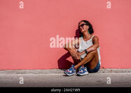 Attraente donna anziana con tintarella indossando occhiali da sole appoggiata contro una brillante luce rosa parete monocromatica. Soleggiata giornata di sole una persona di più femmina matura. Foto Stock