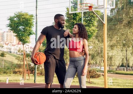 Team perfetto. Coppia africana in piedi con la sfera che abbraccia il campo da pallacanestro guardando ogni altra felice Foto Stock