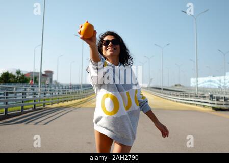 una ragazza in una felpa con cappuccio sportiva lancia un arancio come un baseball. sport, fitness Foto Stock