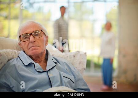 Senior infelice uomo disteso nel letto con sua moglie e un amico in background. Foto Stock