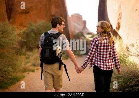 Felice coppia giovane e il loro cane a camminare su un sentiero attraverso le formazioni rocciose nel deserto Foto Stock