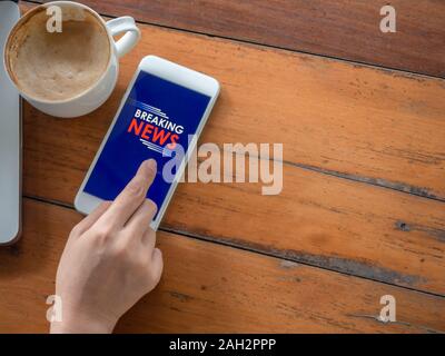 Ultime notizie on line sul concetto dello smartphone. Dito toccando lo schermo dello smartphone su un tavolo di legno con tazza di caffè, vista dall'alto con copia spazio. Foto Stock