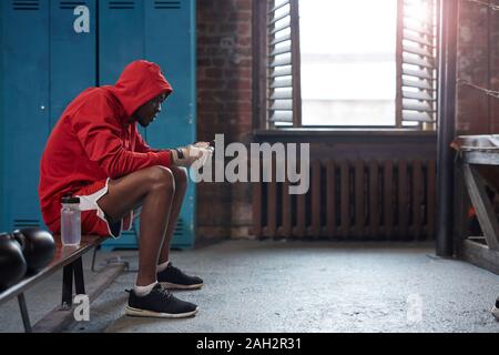 Boxer muscolare in abbigliamento sportivo seduta sul banco di lavoro e utilizzando il suo telefono cellulare in spogliatoio Foto Stock