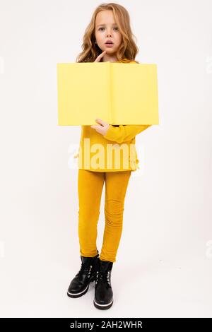 Felice adolescente ragazza con i capelli rossi, felpa con cappuccio e pantaloni gialli letto grande libro giallo isolato su sfondo bianco. Foto Stock