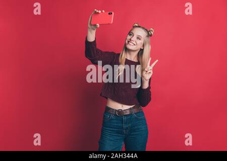 Foto di influencer lady tenendo il telefono in mano rendendo selfies mostra v-simbolo segno di usura del ponticello a maglia e jeans isolato di color pastello di colore rosso Foto Stock