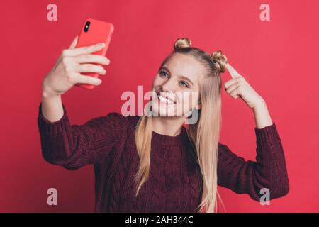 Foto di influencer lady tenendo il telefono in mano rendendo selfies mostrando una nuova acconciatura ai seguaci di usura del ponticello a maglia isolato di color pastello di colore rosso Foto Stock