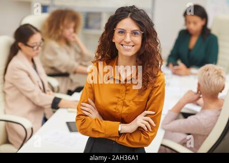 Donna elegante con lunghi capelli ricci in piedi con le braccia chiuse indossando occhiali da vista guardando la telecamera sorridente con i colleghi coworking dietro di lei, media ritratto Foto Stock