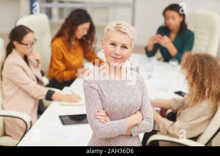 Fiducioso donna matura in piedi con le braccia chiuse guardando fotocamera a sorridere con i colleghi coworking dietro di lei, media ritratto Foto Stock