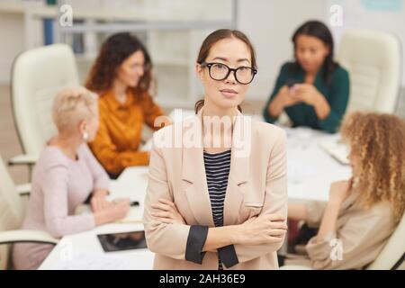 Attraente donna asiatica in piedi con le braccia incrociate guardando la telecamera con i colleghi coworking dietro di lei, alla cintura su ritratto Foto Stock