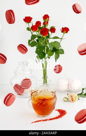 Flying cibo e ancora il concetto di vita. La colazione dessert su sfondo bianco con amaretti, bouquet di rose rosse e spruzzi di tea. Foto Stock