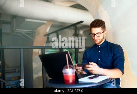 Bello imprenditore distratti dal lavoro sul portatile per guardare video sullo smartphone. Libero professionista tenendo il telefono cellulare e di navigazione mediante alta velocità 4G Foto Stock