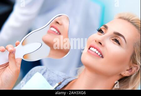 Vicino la fotografia di una donna sorridente in dental clinic. La donna mantiene lo specchio dopo lo sbiancamento dei denti procedura Foto Stock