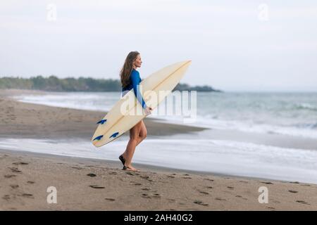 Giovane donna con la tavola da surf in spiaggia, Kedungu Beach, Bali, Indonesia Foto Stock