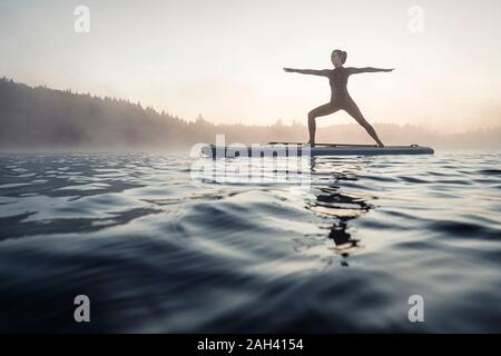 Donna pratica di paddle board yoga sul lago Kirchsee al mattino, Bad Toelz, Baviera, Germania Foto Stock