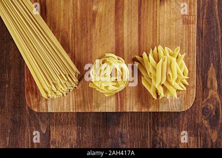 Vari tipi di pasta su tavola di legno con tagliere, vista dall'alto. Pappardelle, spaghetti, Penne su tavola di legno scuro. Foto Stock