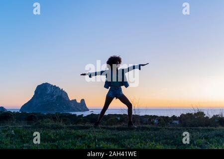 Giovane donna in piedi su un prato a praticare lo Yoga al tramonto, Ibiza Foto Stock