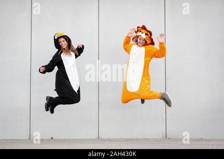 Due donne in penguin e lion costume jumping nella parte anteriore della parete di cemento Foto Stock