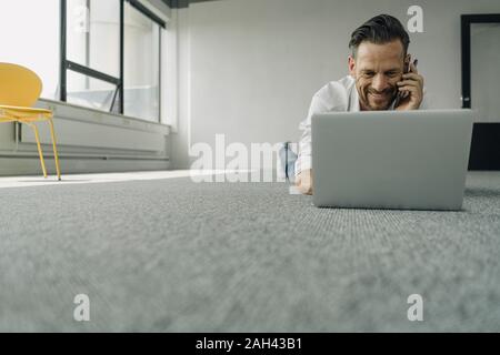 Sorridente imprenditore maturo sdraiato sul pavimento in ufficio vuoto utilizzando computer portatile e lo smartphone Foto Stock