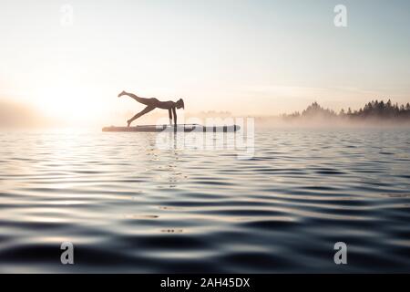 Donna pratica di paddle board yoga sul lago Kirchsee al mattino, Bad Toelz, Baviera, Germania Foto Stock