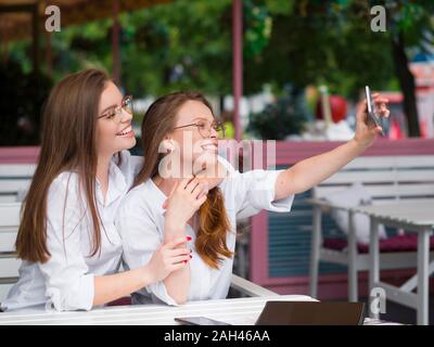 Due ragazze carine in occhiali makeing un selfie seduti in una caffetteria estiva. Reti sociali di concetto Foto Stock