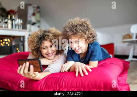 Madre e figlio guardando un video sullo smartphone, giacente sul cuscino di grandi dimensioni Foto Stock