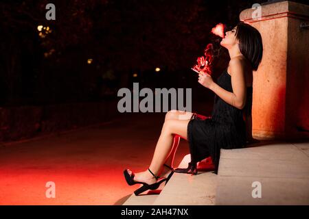 Il fumo giovane donna vestita di nero abito da sera seduti sulle scale di notte Foto Stock
