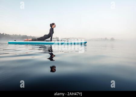 Donna pratica di paddle board yoga sul lago Kirchsee al mattino, Bad Toelz, Baviera, Germania Foto Stock