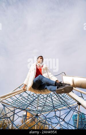 Giovane uomo seduto sul telaio di arrampicata su un parco giochi Foto Stock