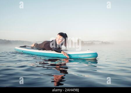 Donna sdraiata sul sup gonfiabile paddleboard sul lago Kirchsee, Bad Toelz, Baviera, Germania Foto Stock