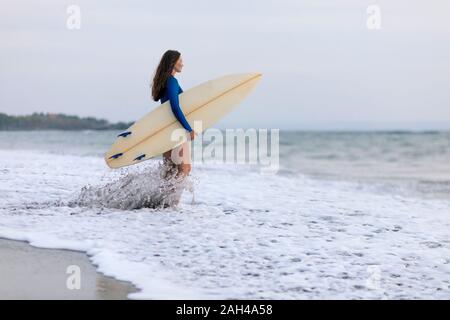 Giovane donna con la tavola da surf in spiaggia, Kedungu Beach, Bali, Indonesia Foto Stock