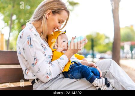 Bottiglia di madre-bambino alimentazione ragazzo su una panchina nel parco Foto Stock