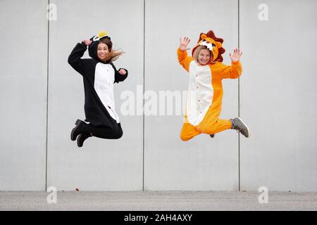 Due donne in penguin e lion costume jumping nella parte anteriore della parete di cemento Foto Stock