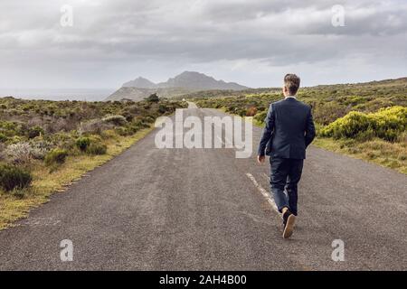 Vista posteriore di imprenditore a camminare sulla strada di campagna, a Cape Point Western Cape, Sud Africa Foto Stock