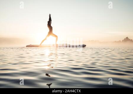 Donna pratica di paddle board yoga sul lago Kirchsee al mattino, Bad Toelz, Baviera, Germania Foto Stock