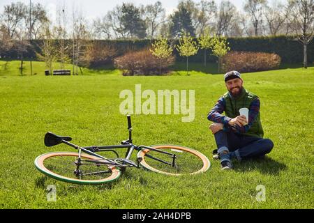Giovane uomo sul prato con una bicicletta. Egli tiene un bicchiere di caffè in mano. Tempo libero in città Foto Stock