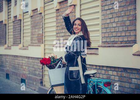 Bella giovane donna sorridente camminando con la bicicletta e agitando la mano sulla strada della citta'. Bellezza,gesto e concetto di stile di vita Foto Stock