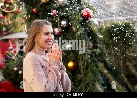 Felice ragazza ridere e gode della neve durante in piedi sul Nuovo Anno Albero di fondo durante il festival tradizionale viaggio per Natale Foto Stock