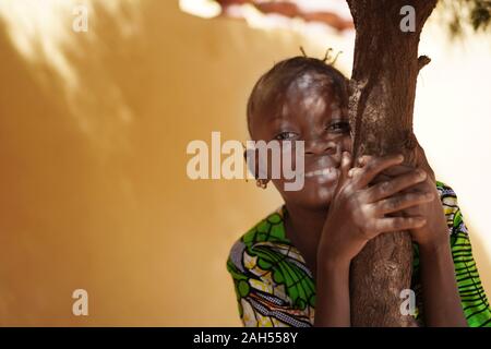 Gli effetti di luce su una sorridente ragazza africana del volto aggrappato ad un tronco di albero Foto Stock