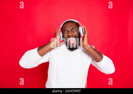 Foto di divertenti pelle scura uomo ascoltare playlist preferita in earflaps, crazy cantando cool via indossare bianco felpa lavorata a maglia isolato sfondo rosso Foto Stock