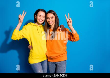 Foto di pretty due amiche lady mostra v-segno abbracciando simboli, indossando luminoso casual felpe con cappuccio e jeans isolato vibrante blu colore di sfondo Foto Stock