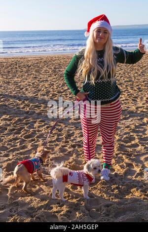 Boscombe, Bournemouth, Dorset, Regno Unito. Giorno di Natale 25 dicembre 2019. Coraggiosi volontari tuffarsi in mare per una nuotata o un tuffo in una bella giornata di sole, per l'annuale beneficenza White Christmas DIP, il più grande tuffo di Natale nel Regno Unito, vestito con abiti eleganti e raccogliere soldi per Macmillan cura locale a Christchurch, Un'unità di assistenza palliativa specialistica per i pazienti della comunità locale. Più di mille prendono parte all'evento che è diventato una tradizione popolare per molti prima del loro pranzo di Natale. Giovane donna con cani nei loro cappotti di Natale. Credit: Carolyn Jenkins/Alamy Live News Foto Stock