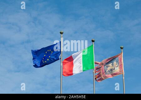 Tre bandiere con il leone di Venezia, l'Italia e la Ue o Unione europea battenti affiancati da pennoni contro un cielo blu con spazio di copia Foto Stock
