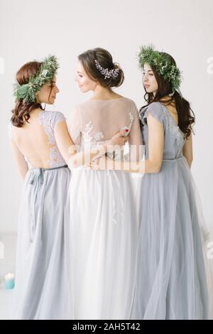 Nozze. Bella sposa in un abito bianco in piedi e abbracciando bridesmaids in grigio blu abiti e ghirlande. Foto Stock