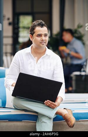 Ritratto di indiani positivo uomo seduto su un banco di lavoro con computer portatile aperto e pensando a una soluzione per il suo progetto Foto Stock