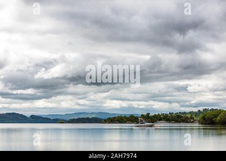 Un lago scozzese circondato da alberi e vegetazione, con un motoscafo ormeggiato in mezzo e lontane montagne sotto un cielo nuvoloso Foto Stock