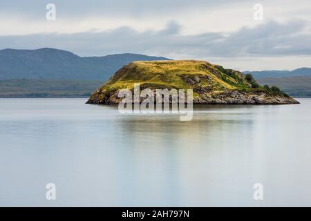 Una piccola isola rocciosa coperta di erba verde in un lago scozzese con montagne lontane Foto Stock