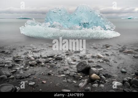 Primo piano di una spiaggia islandese con sabbia nera e ciottoli in primo piano, e un piccolo iceberg e alcune onde infrangenti sullo sfondo Foto Stock